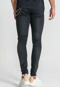 Jeans skinny negros con una textura suave, con un adorno de cadena en el bolsillo trasero y un diseño ajustado. Combinados con zapatillas blancas y beige.