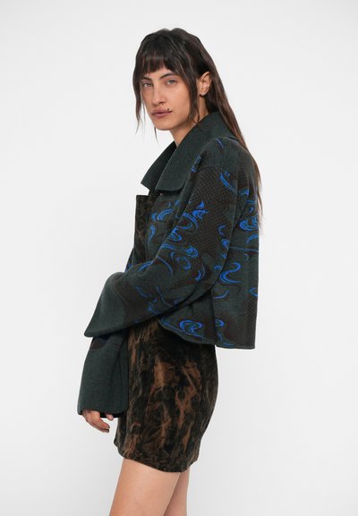 Casaco verde com padrão e detalhes em azul, colarinho oversized, combinado com uma saia de cortiça castanha. Projetado com materiais texturizados.
