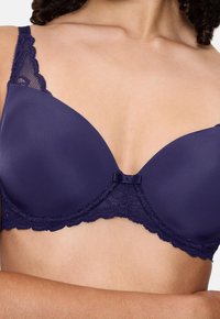 Navy modra bralette ima gladke skodelice, čipkaste detajle na naramnicah in pasu ter majhno pentljo na sredini za dodano okrasitev.