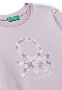 Lavendelfarbenes Rundhals-Shirt mit floral-musterndem Benetton-Logo in weißem und lila Glitzer auf der Vorderseite sowie grünem Markenlabel am Kragen.