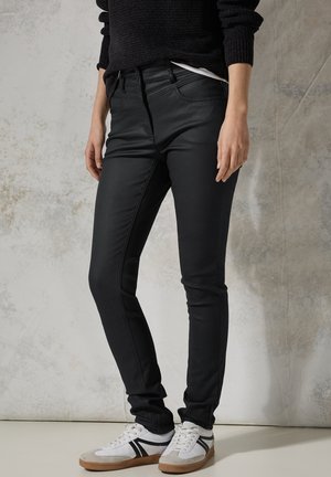 Zwarte, slim-fit jeans met een gladde textuur, voorzien van zakken aan de voorkant en contrasterend stikwerk, gecombineerd met witte sportschoenen en zwarte accenten.