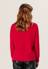 Roter Wollpullover mit rundem Ausschnitt, geripptem Saum und langen Ärmeln, kombiniert mit schwarzen Lederhosen. Glatte Textur, schlichtes Design.