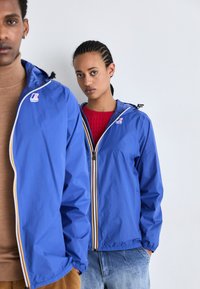 Chaquetas azules resistentes al agua con cremalleras frontales y capuchas, que presentan rayas multicolores y acentos de logo, llevadas por dos modelos.