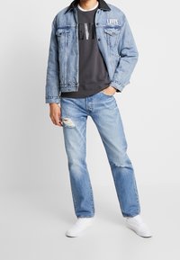 Hombre vistiendo una chaqueta de mezclilla Levi's azul claro y jeans, con una camiseta negra de Levi's y zapatillas blancas, de pie con una mano en el bolsillo.