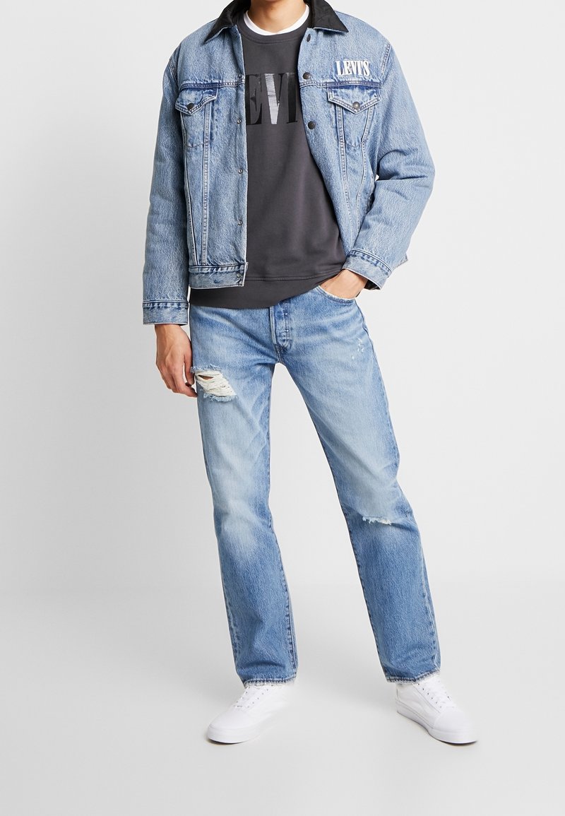 Hombre vistiendo una chaqueta de mezclilla Levi's azul claro y jeans, con una camiseta negra de Levi's y zapatillas blancas, de pie con una mano en el bolsillo.