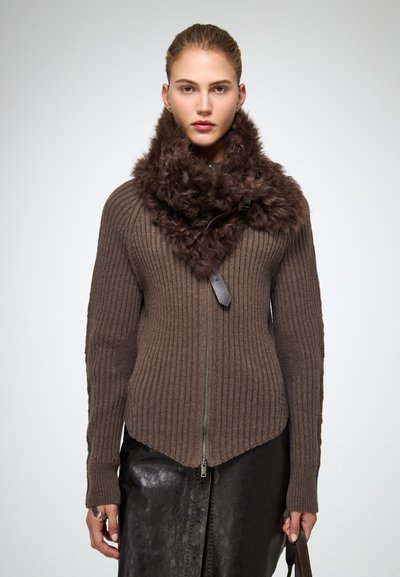 Donna che indossa un maglione a costine marrone con una grande collare in pelliccia marrone scuro e una gonna in pelle nera, mentre tiene il manico di una borsa marrone.