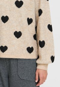 Cremefarbener Pullover mit schwarzen Herzmotiven, gerippten Bündchen und strukturiertem Finish. Kombiniert mit grauen, strukturierten Stoffhosen.