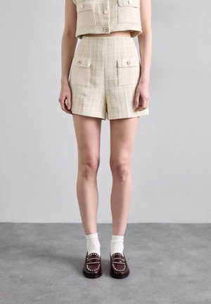 Beige ja keltaiset tweed-shortsit, joissa on kaksi etutaskua ja lyhyt yhteensopiva liivi, jota korostavat vaaleanpunaiset napit. Mallilla on valkoiset sukat ja loaferit.