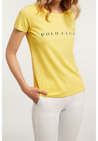 Żółta bawełniana koszulka z okrągłym dekoltem i krótkimi rękawami, z napisem „POLO CLUB” w ciemnozielonym kolorze na wysokości klatki piersiowej. W połączeniu z białymi spodniami.
