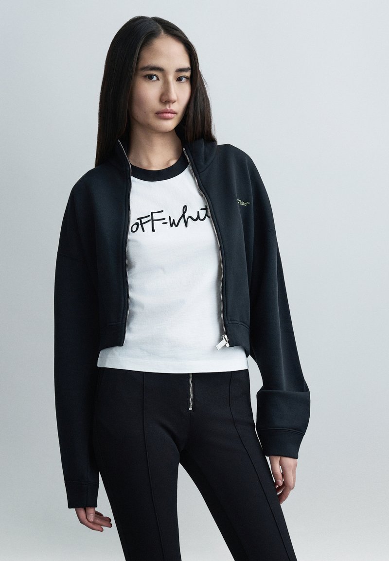 Κοντό μαύρο φούτερ με φερμουάρ και φαρδιά μανίκι α, φορεμένο πάνω από λευκό t-shirt με μαύρο κείμενο σε γραφή. Μαύρο εφαρμοστό παντελόνι με λεπτομέρεια φερμουάρ.