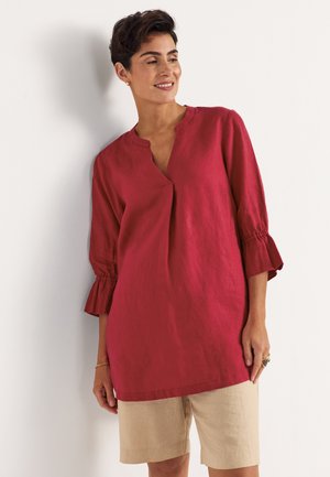 Uśmiechnięta kobieta z krótkimi ciemnymi włosami, ubrana w czerwoną tunikę z długim rękawem i beżowe szorty do kolan, opierająca się o białą ścianę.