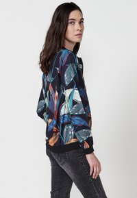 Giacca bomber floreale in blu scuro con vivace stampa di foglie tropicali. Polsini e orlo a costine per un migliore fitting. Indossata con jeans scuri e texturizzati.