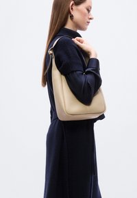 Μπεζ δερμάτινη τσάντα ώμου με ανώμαλη υφή, στρογγυλεμένες άκρες και έναν ενιαίο ιμάντα ώμου. Το μοντέλο φοράει μπλε σκούρο φόρεμα-πουλόβερ.