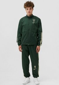 Tuta Nike verde con strisce crema, con logo sulla giacca e sui pantaloni. Realizzato in materiale leggero, progettato per abbigliamento sportivo.