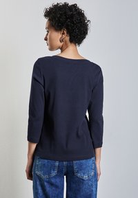 Marineblauer gerippter Pullover mit Dreiviertelärmeln, ausgestattet mit einem weiten Ausschnitt und glatter Textur, kombiniert mit hellblauen Denim-Jeans.