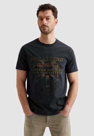 Man met kort bruin haar en baard, gekleed in een zwart "PME Legend" grafisch T-shirt en beige broek, met handen in de zakken, tegen een effen achtergrond.
