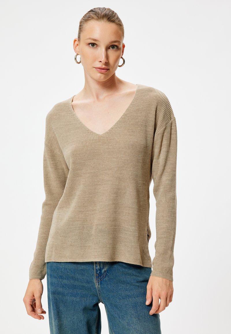 Beige gestrickter Pullover mit V-Ausschnitt, langen Ärmeln und lockerer Passform. Die Textur ist gerippt und wird mit blauen hochtaillierten Jeans kombiniert.