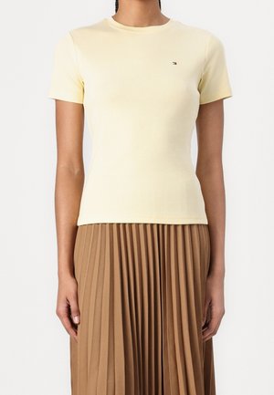 Kobieta w dopasowanym, jasnopopielatym krótkim topie z małym logo na piersi oraz w brązowej plisowanej spódnicy z wysokim stanem.