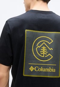 Μαύρο κοντομάνικο μπλουζάκι με μεγάλο χρυσό λογότυπο που απεικονίζει βουνά και δέντρο. Το ύφασμα φαίνεται λείο με διακριτική λάμψη.