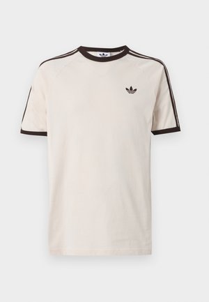 Camiseta beige de Adidas con cuello negro, bordes de las mangas y tres franjas negras a lo largo de los hombros; logotipo de Adidas en el pecho izquierdo.