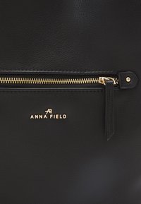 Svart läderväska med en texturerad yta, gulddragkedja och logotypen "ANNA FIELD" i guldbokstäver på framsidan.