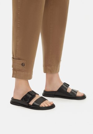 Schwarze Ledersandalen mit verstellbarem Riemen, offenem Zehendesign und robustem Außenboden, kombiniert mit hochgekrempelten hellbraunen Hosen.