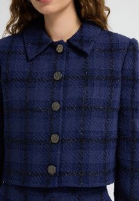Marinblå rutig jacka i texturerat tyg med krage, prydd med sex runda, mörka knappar och metalltrådsdetaljer.