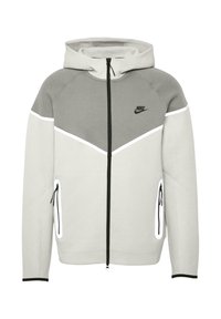 Sudadera con cremallera Nike en gris y blanco, con capucha, que presenta un diseño en forma de chevrón, bolsillos laterales con cremalleras y detalles en negro. Tela elástica.