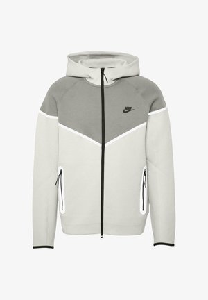 Hallide ja valge Nike kapuutsiga tõmmatav sviiter, millel on chevroni disain, küljetaskud lukuga ja mustad aktsendid. Veniv kangas.