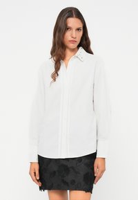 Camicia bianca con colletto texturizzato e dettagli decorativi. Maniche lunghe, taglio slim, tessuto liscio. La gonna presenta un motivo floreale scuro.