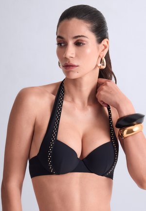 Femme aux cheveux foncés portant un haut à nouer noir clouté, de grandes boucles d’oreilles dorées et de larges bracelets noirs et dorés, regardant vers sa gauche.