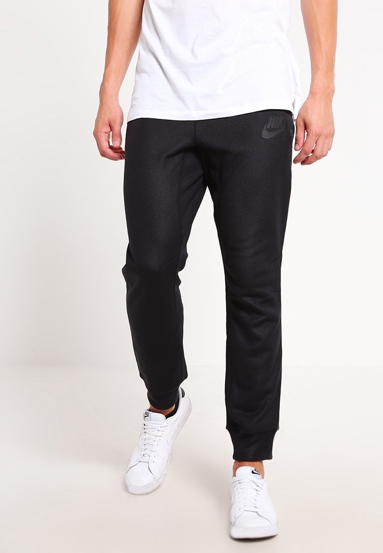 Man i svarta Nike joggarbyxor, vita Nike-sneakers och en vit t-shirt, stående framför en neutral bakgrund.