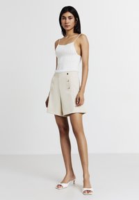 Femme debout, portant un haut à manches sans épaules côtelé blanc, un short taille haute beige avec des boutons, et des sandales à talons ouverts blanches.