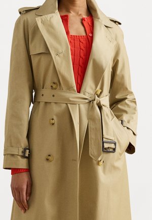 Tan dubbele rij knoop trenchcoat met riem gestrikt in de taille, gedragen over een rood kabelgebreide trui met knoopsluiting bij de hals.