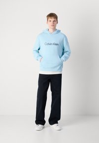Svetlo modrá mikina s kapucňou a prednou vreckom vo forme kengury, s čiernym logom "Calvin Klein". V kombinácii s tmavými nohavicami a bielymi teniskami.