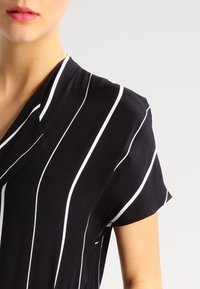 Černý oděv s bílými svislými pruhy, krátkými rukávy a výstřihem do V. Tkanina vypadá lehce a hladce na dotek.