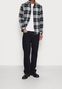 Černé a šedé kárované flanelové tričko přes bílé tričko, spárované s volnými černými kalhotami a černými botami, stojící na neutrálním pozadí.