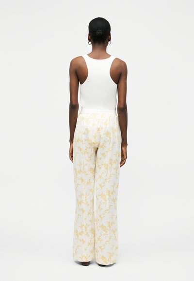 Γυναίκα με σκούρο δέρμα και μαλλιά σε κότσο, που φοράει λευκό αμάνικο ριμπ πλεκτό τοπ και λευκό φαρδύ παντελόνι με ανοιχτό κίτρινο λουλουδένιο σχέδιο, όρθια με την πλάτη προς τα πίσω.