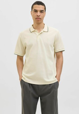 Polo en color beige claro con un acabado texturizado, presenta un cuello con acentos en verde oscuro. Combinado con pantalones oscuros y bien ajustados.
