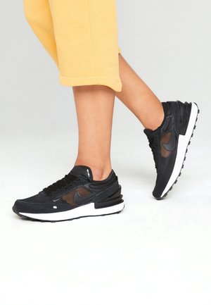 Black Nike sneakers feature a suede and mesh upper, a cushioned white midsole, and a distinct textured black outsole.