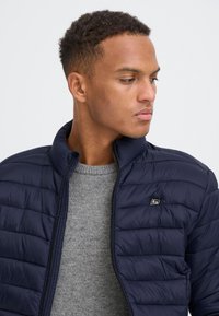 Ung mand kigger mod højre, iført en mørkeblå quiltet jakke over en grå strikket sweater, mod en ensfarvet lys baggrund.