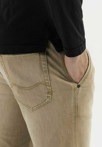 Khaki-Jeans mit einer strukturierten Oberfläche, ausgestattet mit einer Seitentasche und kontrastierenden Nähten. Kombiniert mit einem schwarzen Langarmshirt.