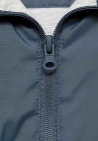 Temno siva zadrga na tkanini, ki ima teksturirane robove vzdolž središča. Gladek povlečni trak z zasnovo z zanko, kontrastni šiv.