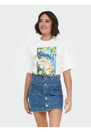 Jeune femme portant un t-shirt blanc avec un imprimé de la côte amalfitaine et une jupe en jean bleu à boutons, souriant légèrement sur un fond uni.