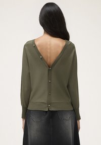 Pulover tricotat de culoare verde măsliniu, cu decolteu adânc în formă de V pe spate, mâneci lungi și detalii cu nasturi de-a lungul coloanei vertebrale. Asortat cu o fustă din denim închis.