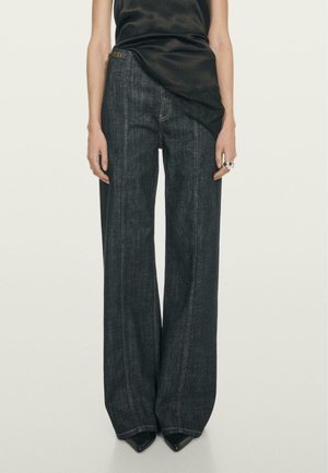 Wide-leg dark denim jeans with a high waist, subtle stitching details, and a smooth texture, paired with pointed black heels.
