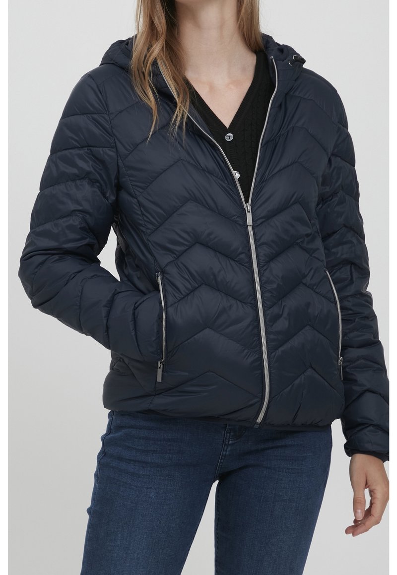 Femme portant une veste matelassée bleu marine à capuche avec fermeture éclair, les mains dans les poches, associée à un jean bleu foncé et un haut noir en dessous.