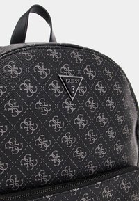 Mochila negra texturizada con un patrón geométrico en gris y una placa central con el logotipo "GUESS" en un triángulo. Cierre con cremallera en la parte inferior.