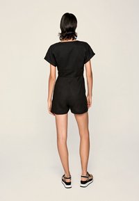 Czarny kombinezon z krótkim rękawem, dopasowaną talią, prostymi spodniami szortami i prostym designem. Tło jest neutralne, beżowe. Buty nie są widoczne.