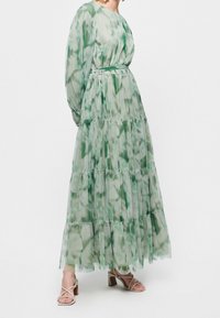 Woman wearing a long, flowy green and white patterned dress with long sleeves and strappy beige block-heeled sandals.